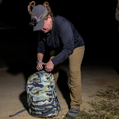 Under the starry night, a person adjusts their headlamp while meticulously packing their durable GR1 USA - Cordura by GORUCK. This rucksack, evocative of the camo backpacks used by Special Forces, stands ready for the adventure ahead.