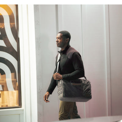 A man strolling outside with a GORUCK Shoulder Bag - Waxed Canvas slung asymmetrically, sporting a black jacket and khaki pants. The shoulder bag features a durable Martexin Wax finish that complements his casual yet polished look.