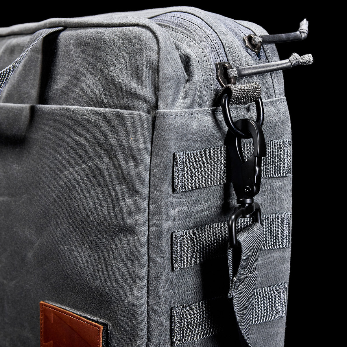 Close-up of a gray fabric bag with a zipper, strap, and metal hook on a black background, showcasing the sleek design of the GORUCK Shoulder Bag - Waxed Canvas, ideal for asymmetrical carries.