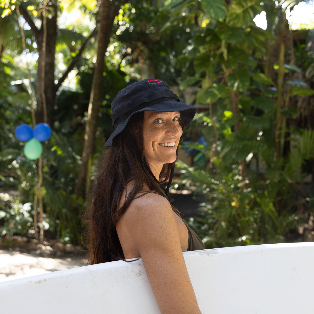 A person with long hair smiles, holding a surfboard in a lush, tropical setting while wearing the Boonie Hat - Slick - ToughDry.