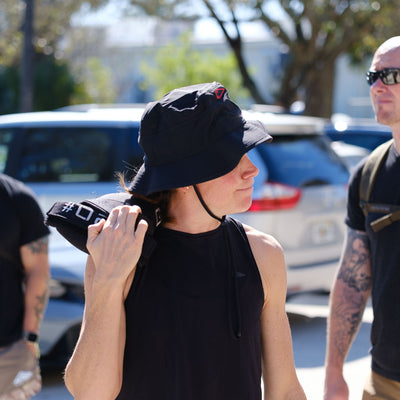 Wearing a Boonie Hat - Slick - ToughDry and a tank top, someone strolls outside, holding a black bag. The sunny day highlights the scene as cars pass in the background, providing shade and protection with each step.