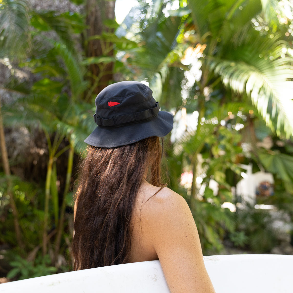 A person with long hair, wearing a Boonie Hat - Slick made from ToughDry material for protection from the elements, stands holding a surfboard among lush tropical plants.