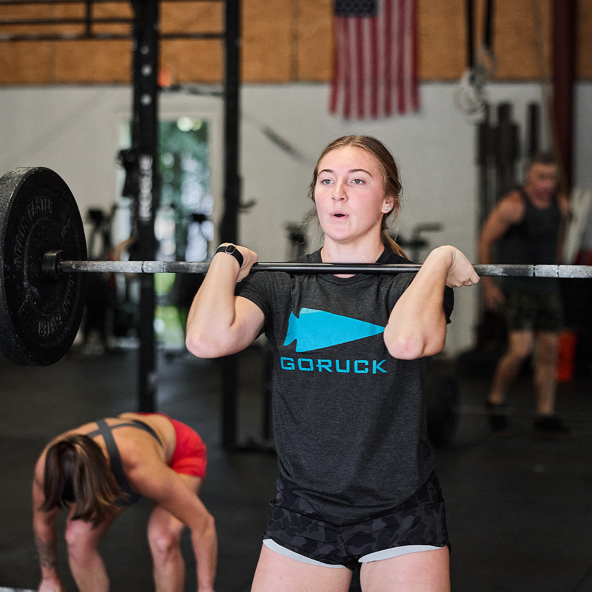 A woman wearing a GORUCK Spearhead Tee - Tri-Blend performs a front squat with a barbell in the gym. An American flag hangs proudly in the background, while another person in athletic wear bends over behind her, reflecting their Special Forces origins.