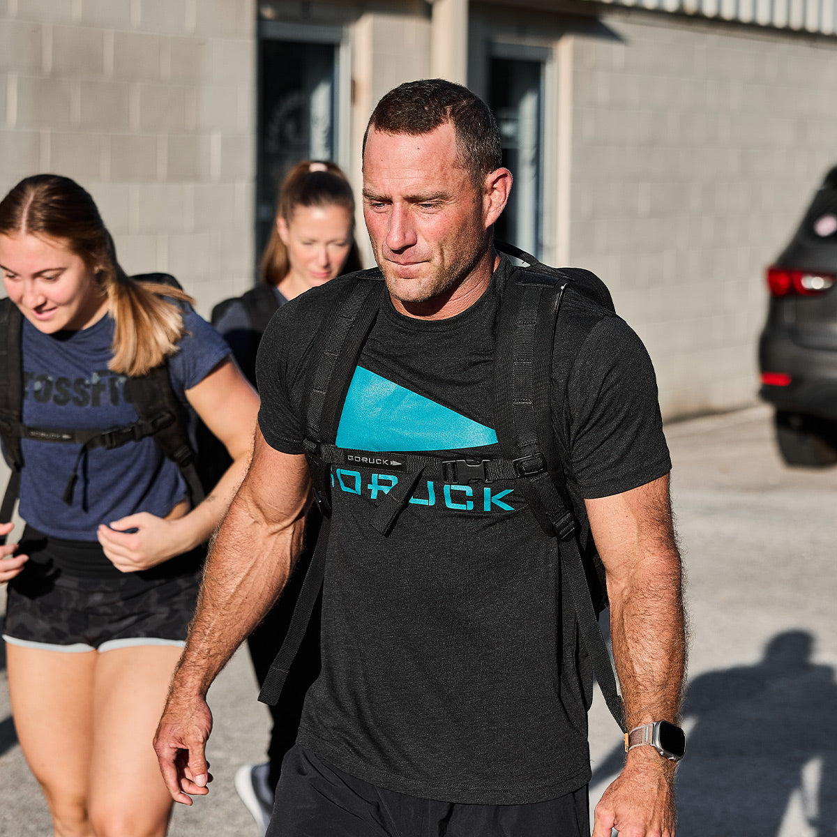 A man wearing a GORUCK Spearhead Tee - Tri-Blend in black, paired with a backpack, strides outdoors under the bright sun. Two women dressed casually and also carrying backpacks follow closely behind him. They walk confidently across a parking lot, truly representing the vendor-unknown brand's GORUCK presence, with a building and car in the background.