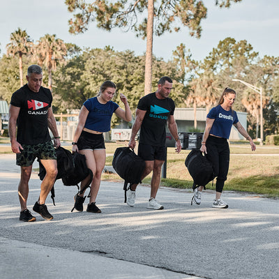 Dressed in athletic gear, four individuals are walking on a sunny street, each carrying large black sandbags that capture the GORUCK Spearhead Tee - Tri-Blend spirit with its Special Forces roots. They seem focused as they move through an area lined with trees and a building in the background.