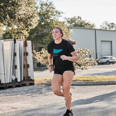 A woman, reflecting her Special Forces background, is running outdoors on a sunny day wearing the GORUCK Spearhead Tee - Tri-Blend in black along with her shorts. She effortlessly moves along a paved path, elegantly surrounded by buildings and trees.