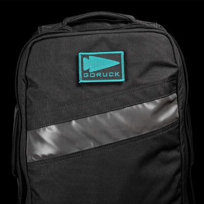 A black backpack featuring a teal GORUCK logo on the upper front and a diagonal reflective strip across the front pocket. It includes an embroidered GORUCK Spearhead patch for added flair, maintaining its subtle, rugged design against the black backdrop.