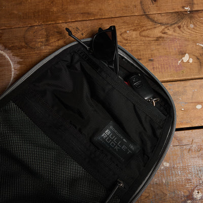 Open the Bullet Ruck Laptop's compartment, where you'll find sunglasses, a car key fob, and a small tag labeled GORUCK 16L. The black mesh and fabric interior, made from durable ROBIC® Ripstop Nylon, is placed on a wooden surface.