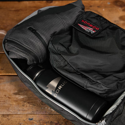 A Bullet Ruck Laptop - Ripstop ROBIC® - 16L from GORUCK sits on a wooden surface, showcasing a rolled-up jacket, folded shirt, and a branded metal water bottle. The partially zipped backpack provides a glimpse of its organized interior and ample lumbar support for enhanced comfort.