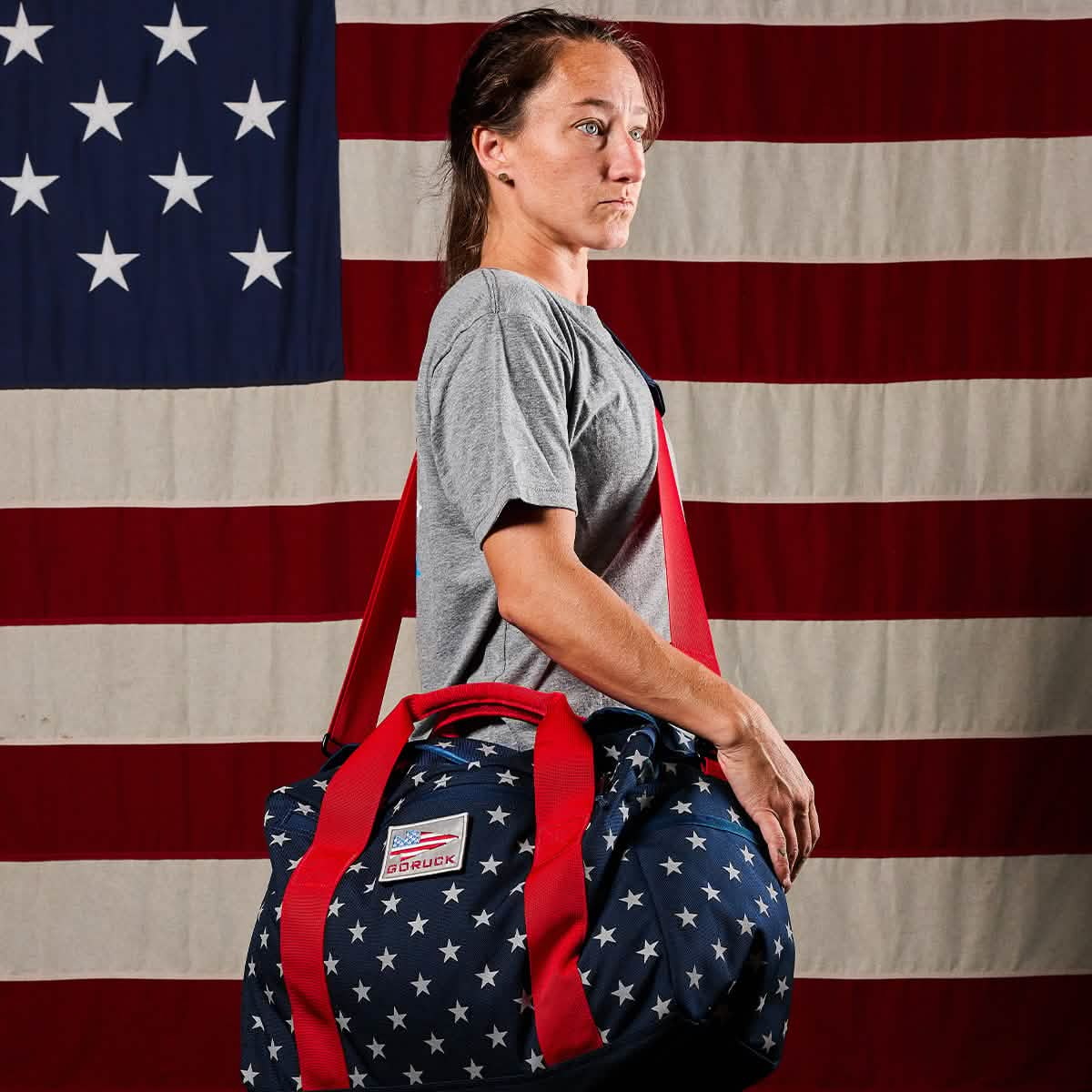 A person proudly stands in front of an American flag backdrop, holding a Kit Bag (Includes Shoulder Strap) adorned with star patterns.