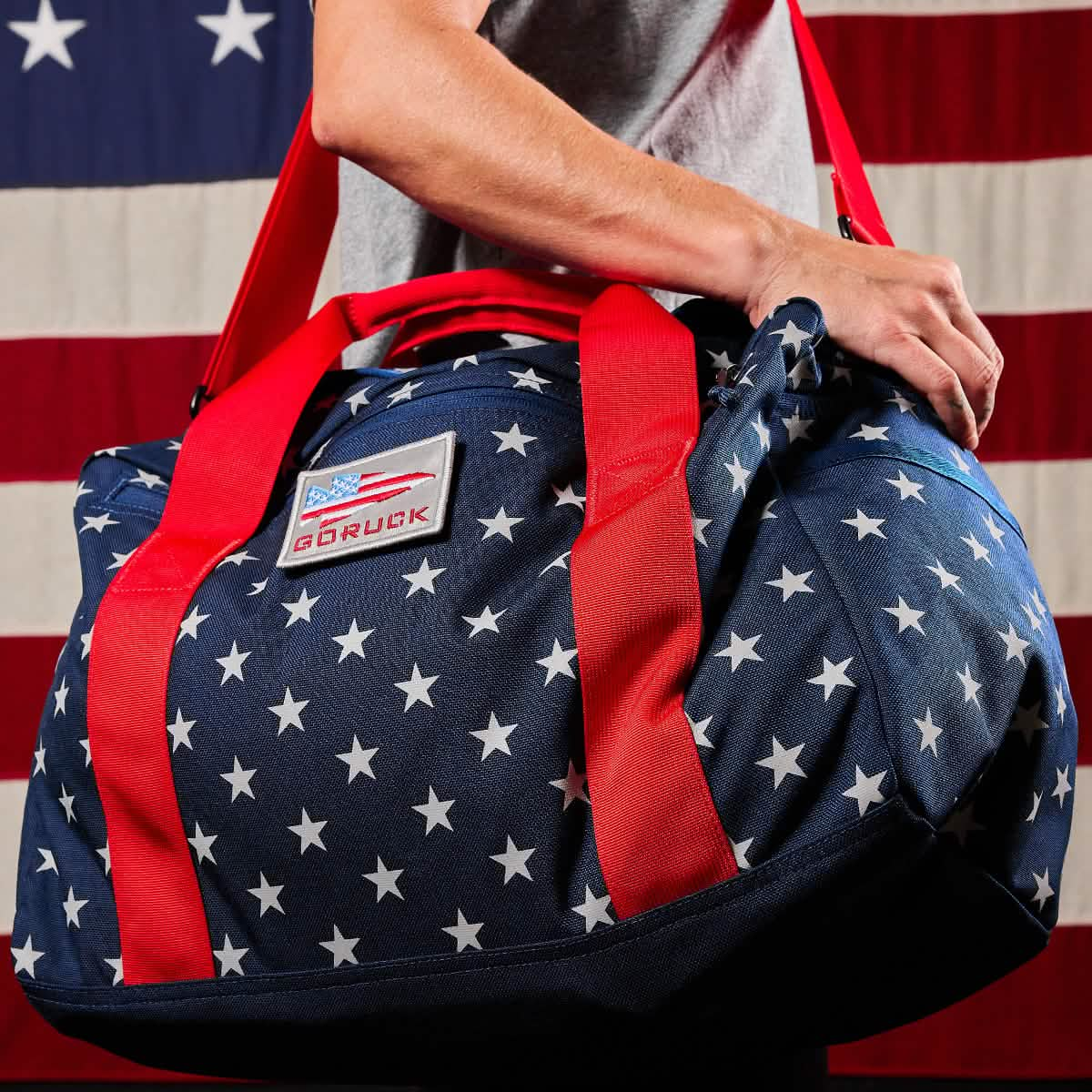 A person holds a Kit Bag (Includes Shoulder Strap) with a star pattern and red straps, reminiscent of an Aviator’s Kit Bag, standing proudly in front of a U.S. flag.