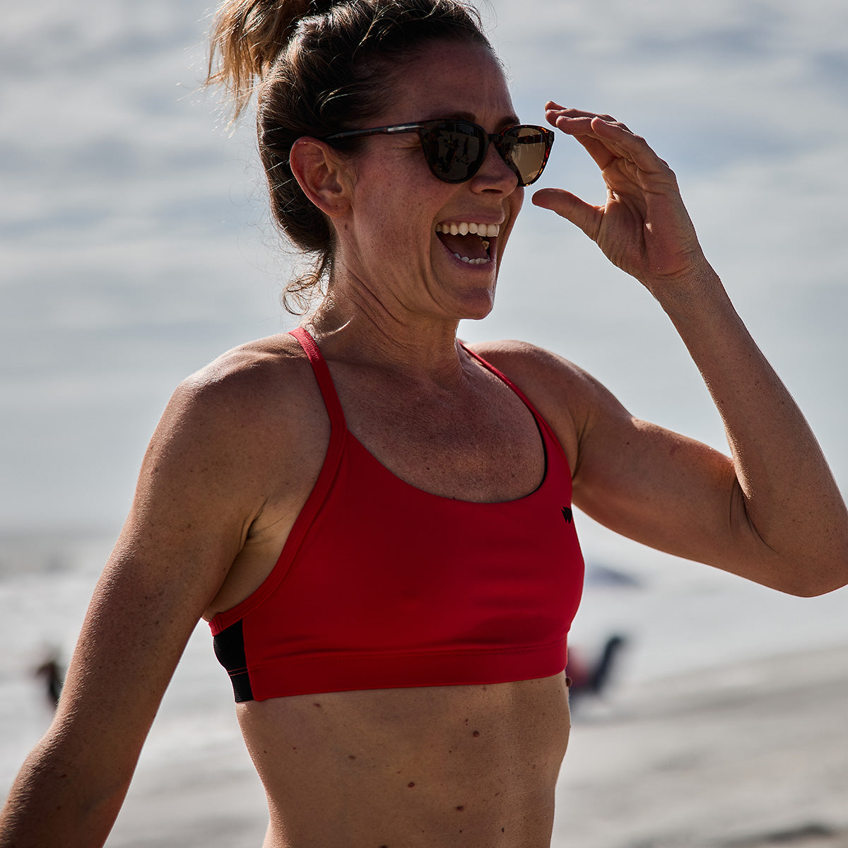 A woman wearing the GORUCK Stealth Bra - ToughFlex in a vibrant red is joyously laughing on a beach, accessorized with sunglasses. The cloudy sky looms above while ocean waves crash in the background.