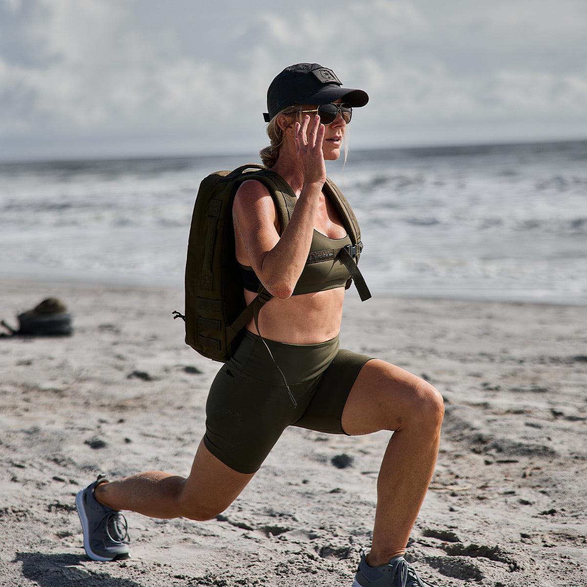 Clad in GORUCK's Stealth Bra - ToughFlex, a person in athletic gear executes a lunge on a sandy beach. The sports bra provides breathable, lightweight support against the backdrop of ocean waves and a cloudy sky.