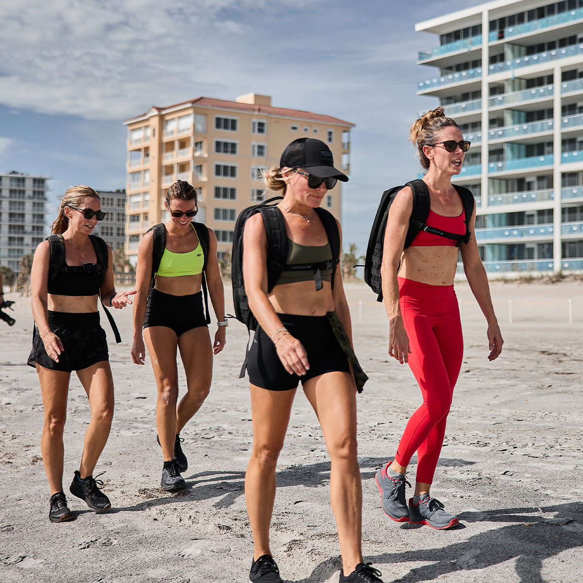 Four women stroll along the sandy beach, sporting athletic gear like the breathable Stealth Bra - ToughFlex by GORUCK and backpacks. The clear sky highlights the residential buildings in the background, making their beach adventure seamless with lightweight support and style.