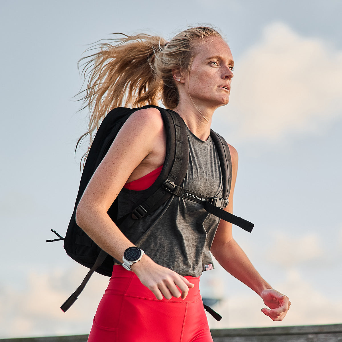 A woman with long blonde hair is running outside, sporting a black backpack and red ToughFlex leggings from GORUCK that provide lightweight support. She pairs these with a gray sleeveless top, and she wears a watch on her left wrist. The sky behind her is partly cloudy.