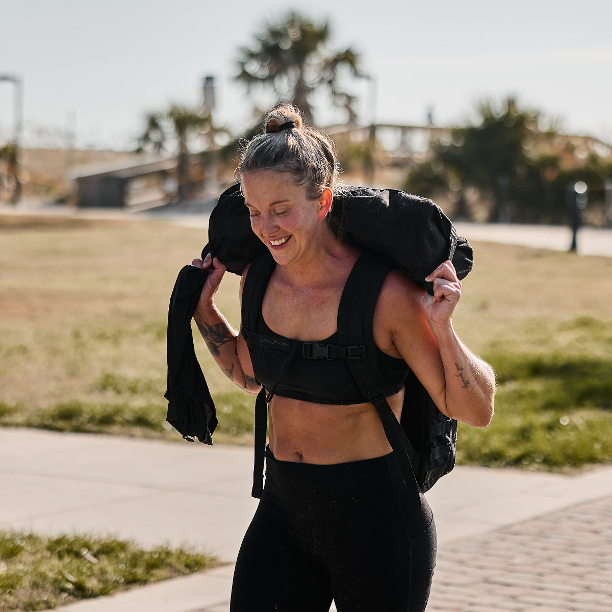 A person in athletic gear is exercising outdoors, carrying a large weighted bag on their shoulders. Dressed in the GORUCK Stealth Bra - ToughFlex, they appear focused and determined, with trees and a park setting in the background.