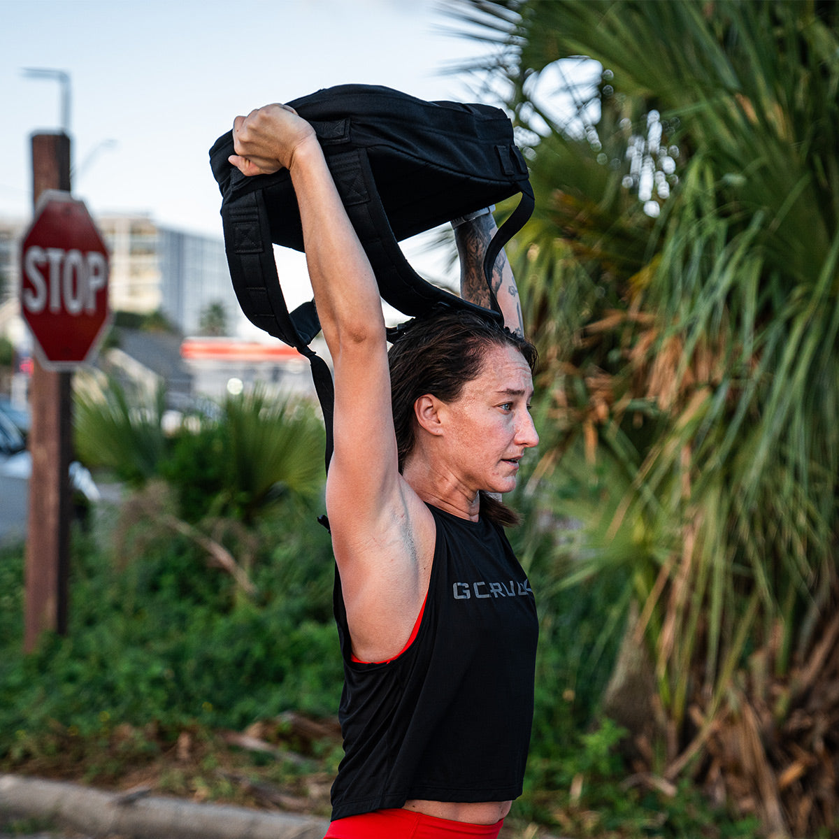 A person exercising outdoors, lifting a backpack overhead, showcases their strength while wearing the Stealth Bra - ToughFlex from GORUCK and red shorts. The scene, featuring a stop sign and palm trees in the background, highlights their use of durable ToughFlex fabric for unparalleled performance.