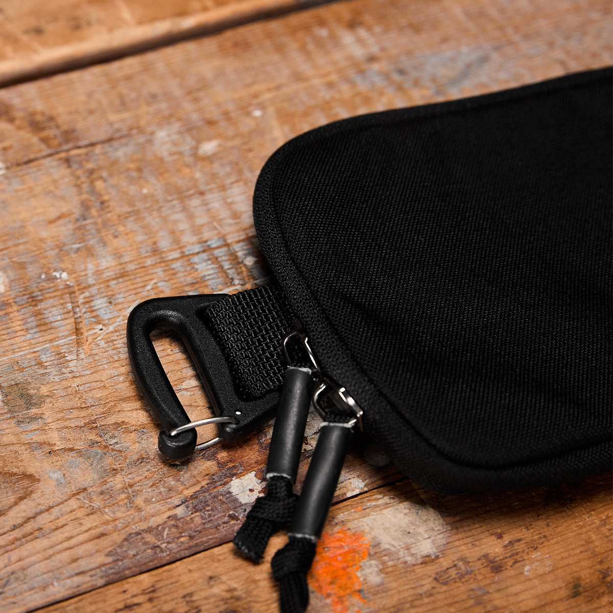 A close-up of the GORUCK Sternum Pouch - Cordura on a wooden surface. The black fabric bag includes a zipper with dual pull tabs and has a black plastic carabiner clip attached to one corner. The worn wood, marked by scratches and a small orange paint swipe, adds character to the scene.