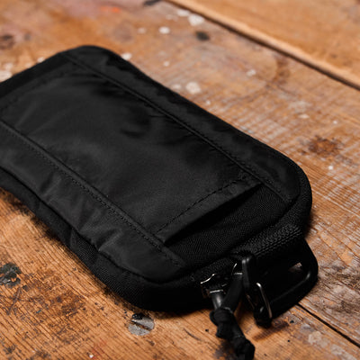 The Sternum Pouch - Cordura by GORUCK is a black zippered pouch with a small carabiner, resting on a rustic wooden surface. It effortlessly integrates into your Rucker gear thanks to its MOLLE-compatible design.