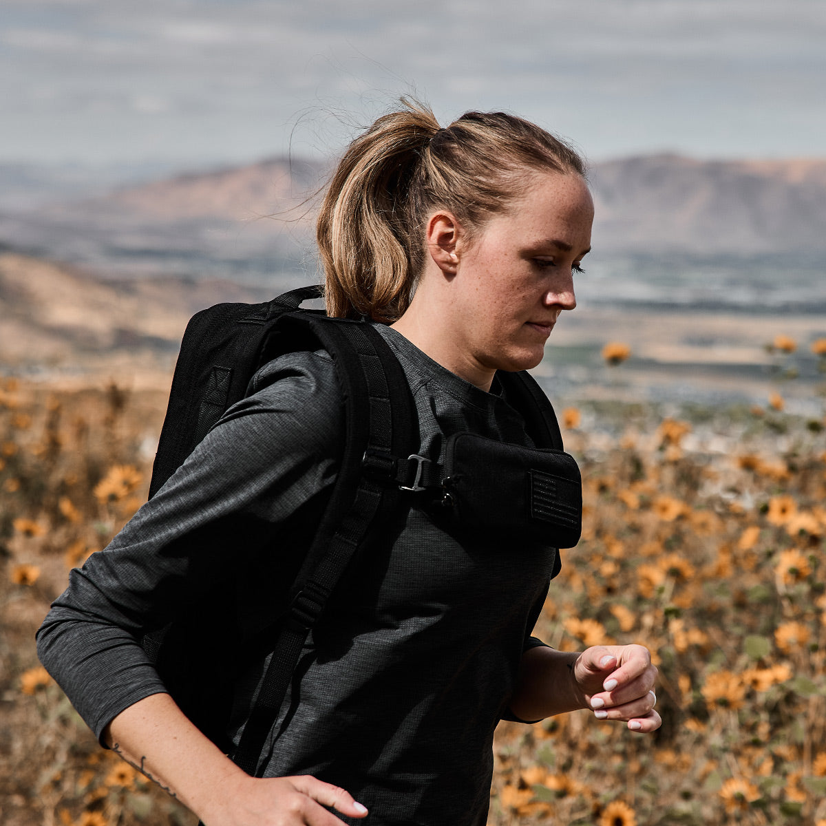 A woman with a ponytail runs outdoors in a dark long-sleeve shirt and a GORUCK Sternum Pouch made from Cordura, worn as a weighted vest. She is surrounded by a picturesque landscape, featuring majestic mountains and vibrant orange wildflowers in the background.