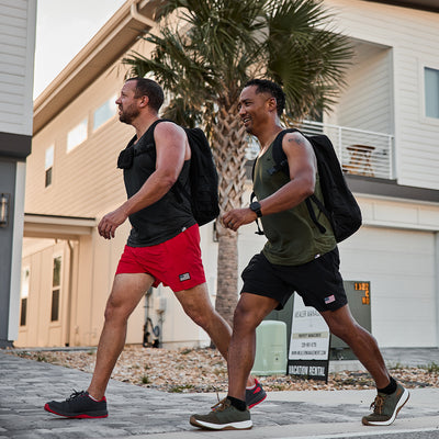 Two men walk down a residential street, each carrying backpacks equipped with MOLLE attachments and Sternum Pouches made from Cordura by GORUCK. They are dressed in sleeveless tops and shorts—one in red, the other in black. Behind them, a palm tree stands tall beside a sign that reads Vacation Rental, while they engage in deep conversation.