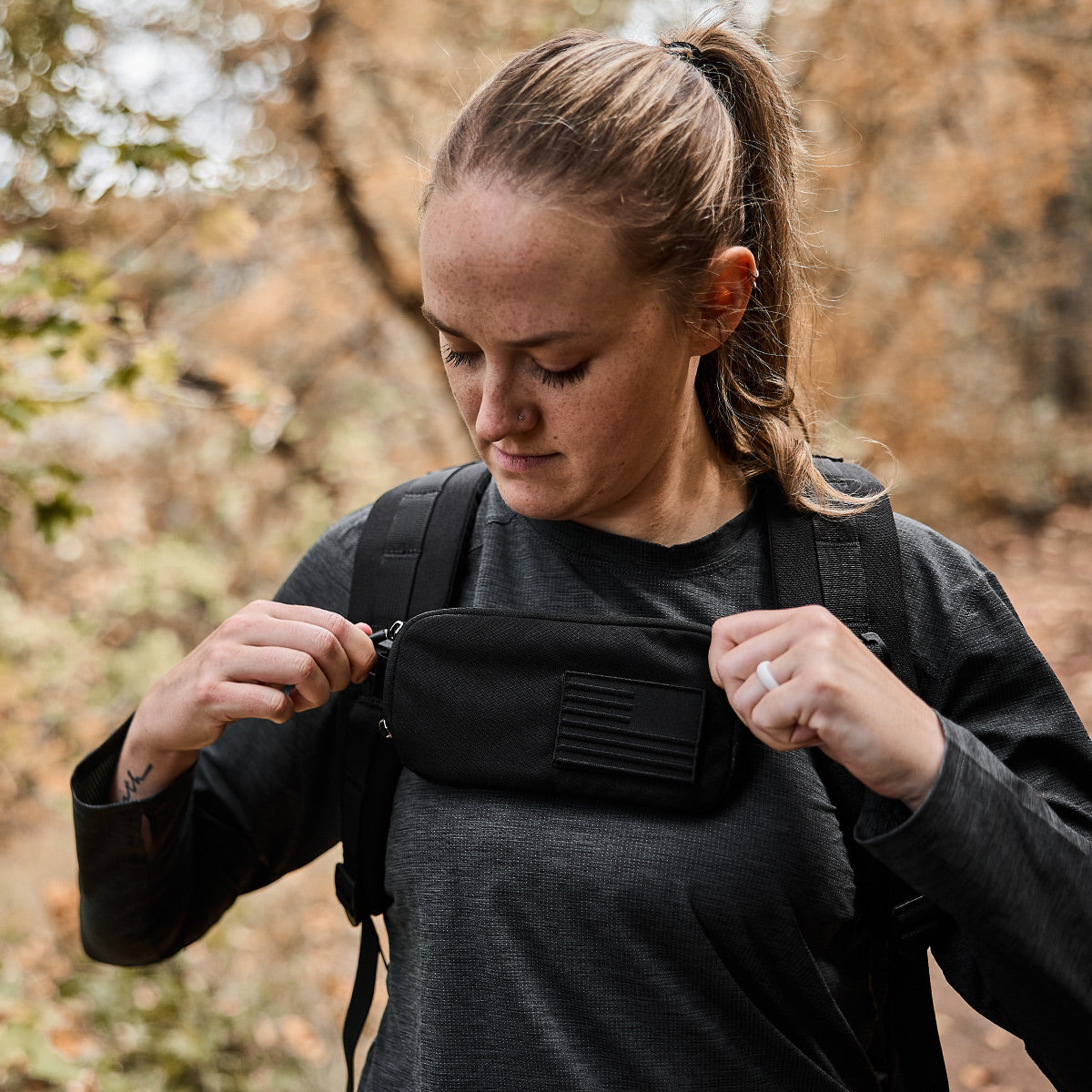 In a forest setting filled with vibrant autumn leaves, a person with a ponytail adjusts a compact black GORUCK Sternum Pouch - Cordura on their backpack. They wear a charcoal gray long-sleeve shirt that is ideal for the crisp autumn weather.