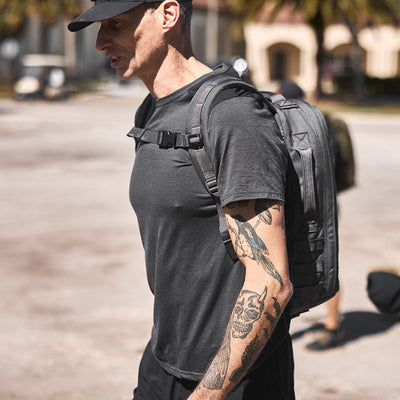 A man with tattoos strolls outdoors, sporting a black cap and T-shirt, along with a GORUCK backpack featuring MOLLE webbing for adjustable weight distribution.