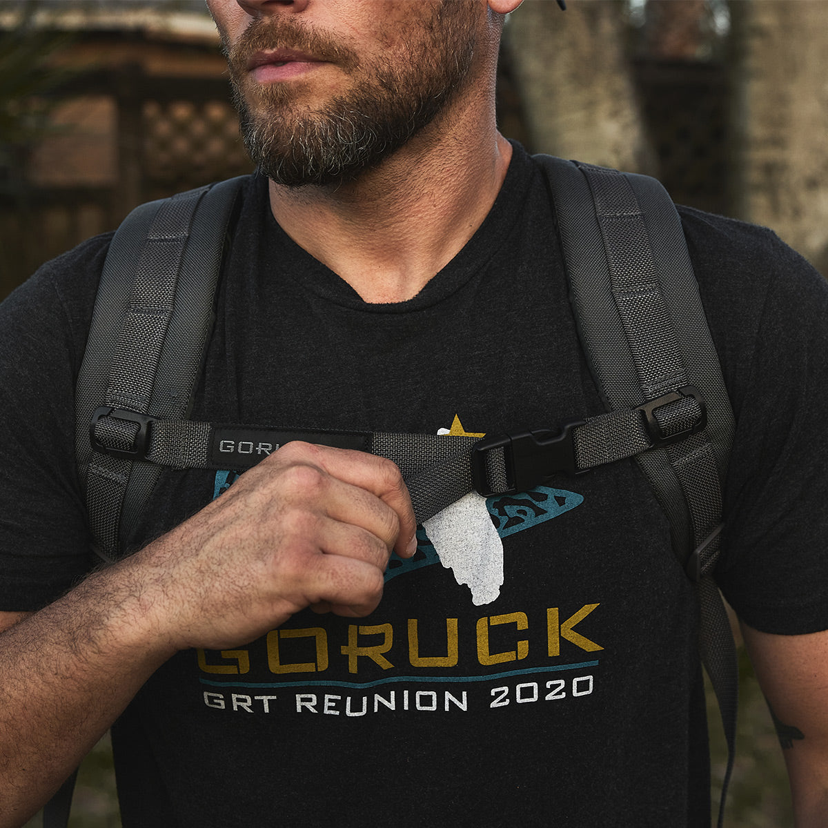 A man with a beard is outdoors wearing a backpack equipped with GORUCK's Training Sternum Strap and a "GORUCK GRT Reunion 2020" shirt.