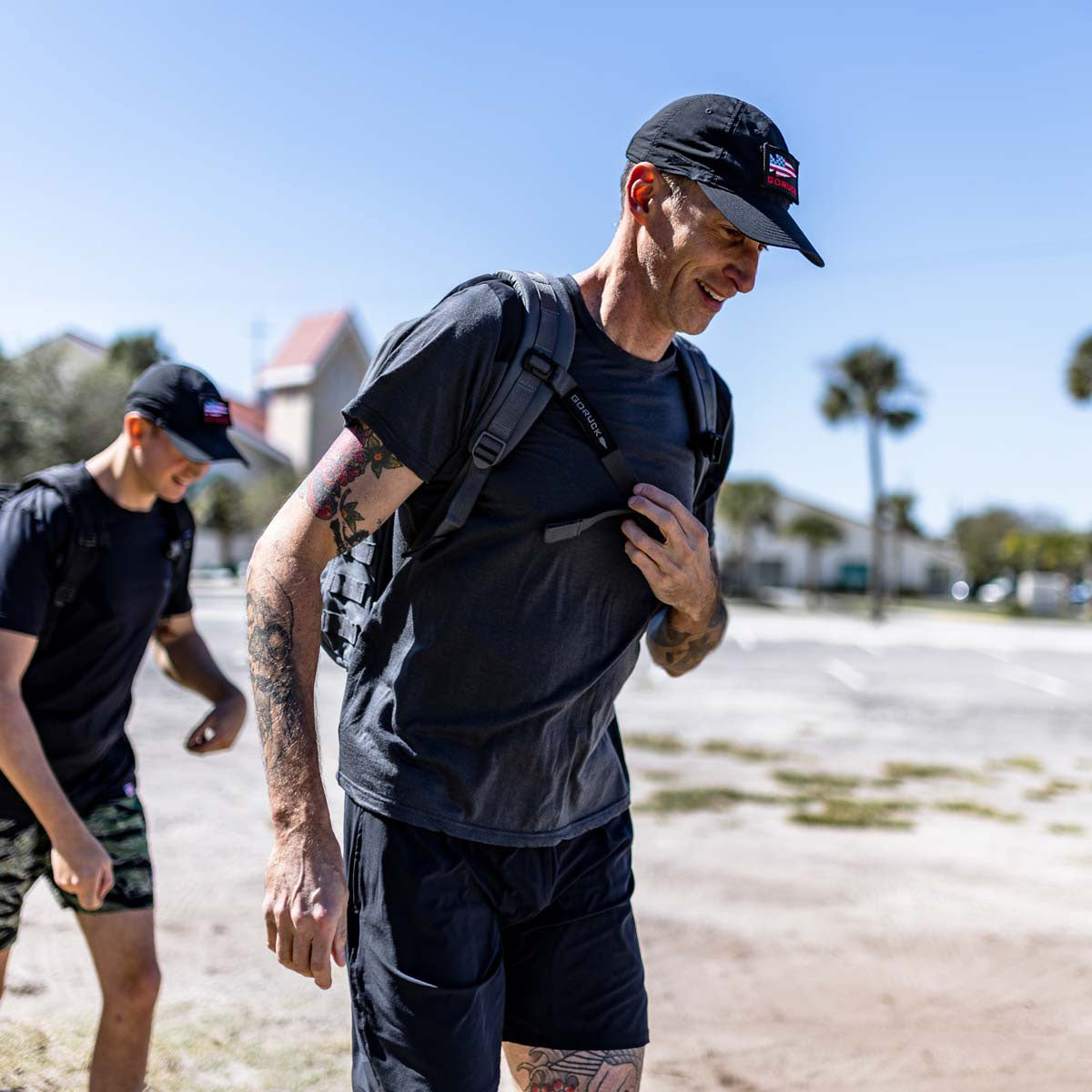 Two individuals in black sportswear and caps are walking outdoors under the sun, featuring their GORUCK Training Sternum Strap which is designed for advanced gear handling with adjustable weight distribution, optimizing comfort during extended hikes.