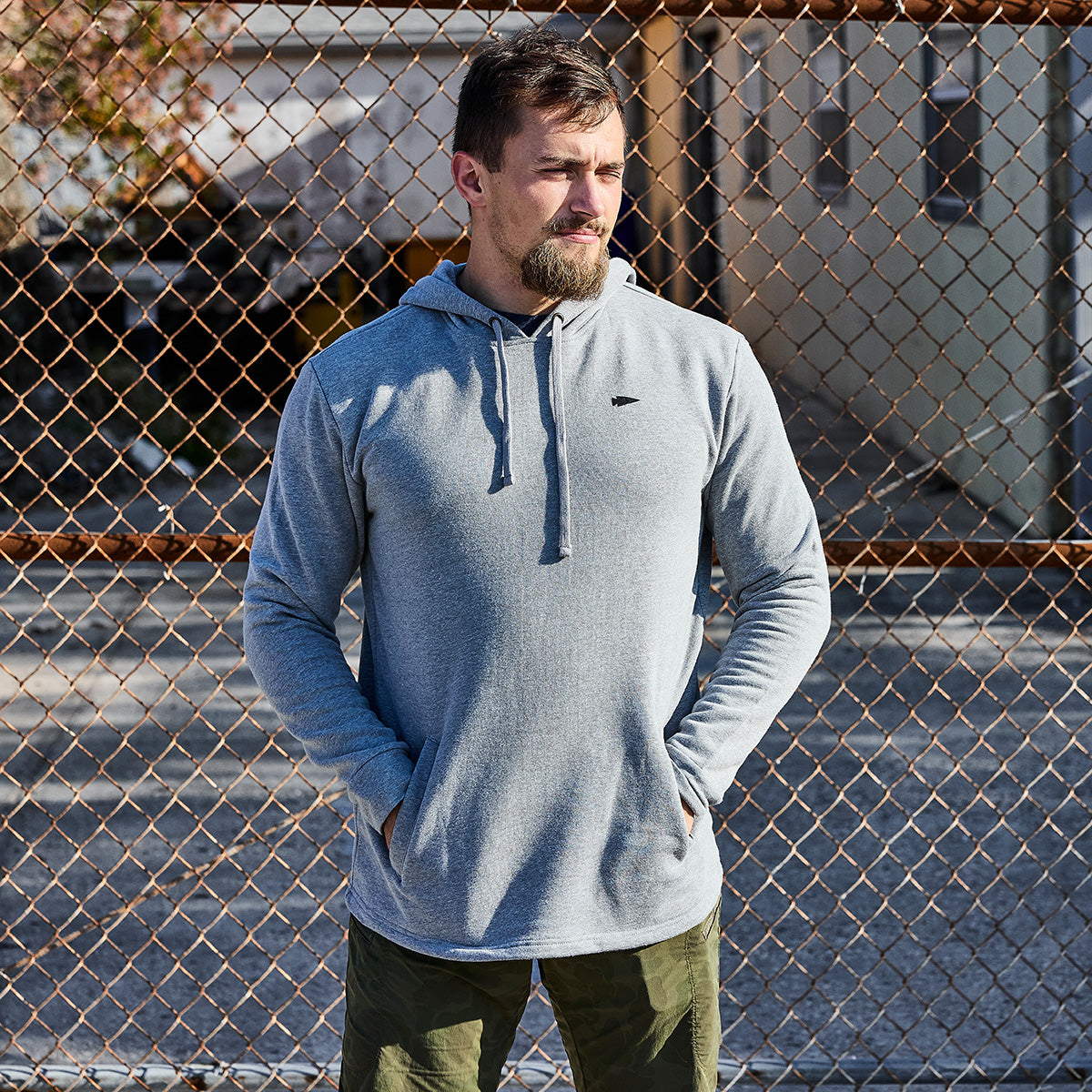 A man in a Subtle Spearhead Hoodie - Tri-Blend and green pants stands confidently before a chain-link fence outdoors, prepared for his rucking adventure.