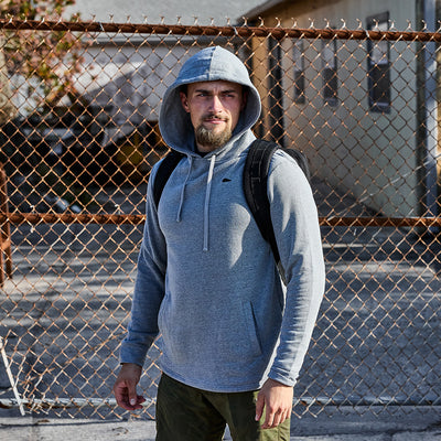 A man in a Subtle Spearhead Tri-Blend Hoodie and backpack stands by a chain-link fence, contemplating the cityscape of buildings in the background.