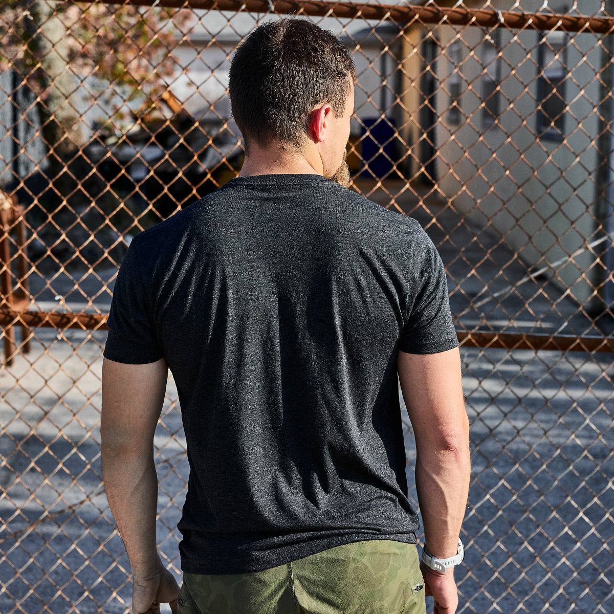 A man with short hair stands facing a chain-link fence, his hands in his pockets. He's wearing a vendor-unknown Subtle Spearhead Tee - Tri-Blend and green camouflage pants, reminiscent of Special Forces attire. The background reveals a paved area and buildings, adding an urban edge to the scene.