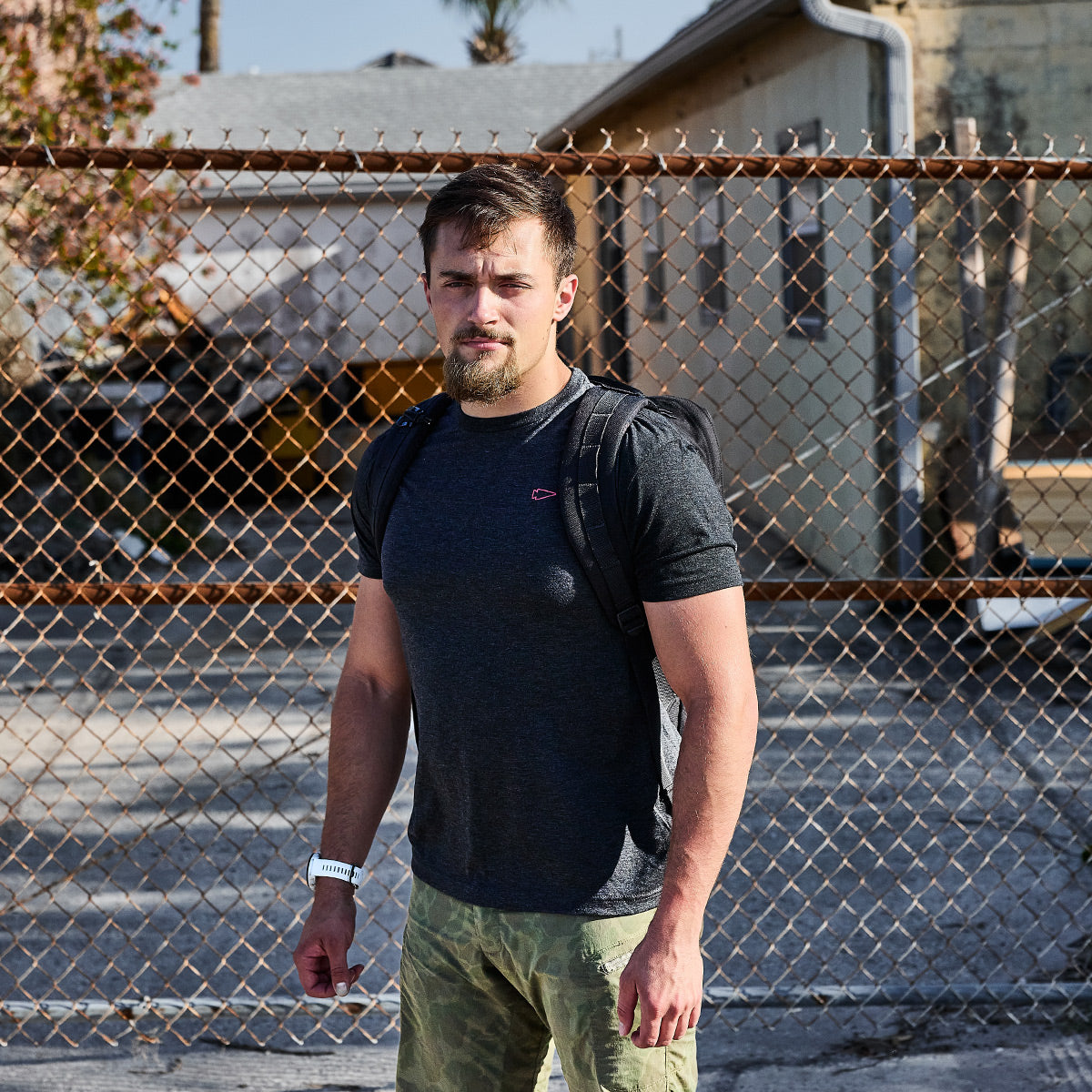 A man with a beard stands in front of a chain-link fence, wearing a Subtle Spearhead Tee - Tri-Blend by vendor-unknown, camo shorts, and a backpack. The setting is outdoors, with buildings and a tree visible in the background.