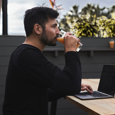 A man wearing the GORUCK Men’s Commando Long Sleeve - Merino Wool shirt sips juice while using his laptop outside.