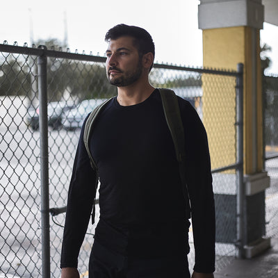 A person wearing a Men’s Commando Long Sleeve made from Merino wool by GORUCK, with a backpack, stands near a chain-link fence under a covered area.