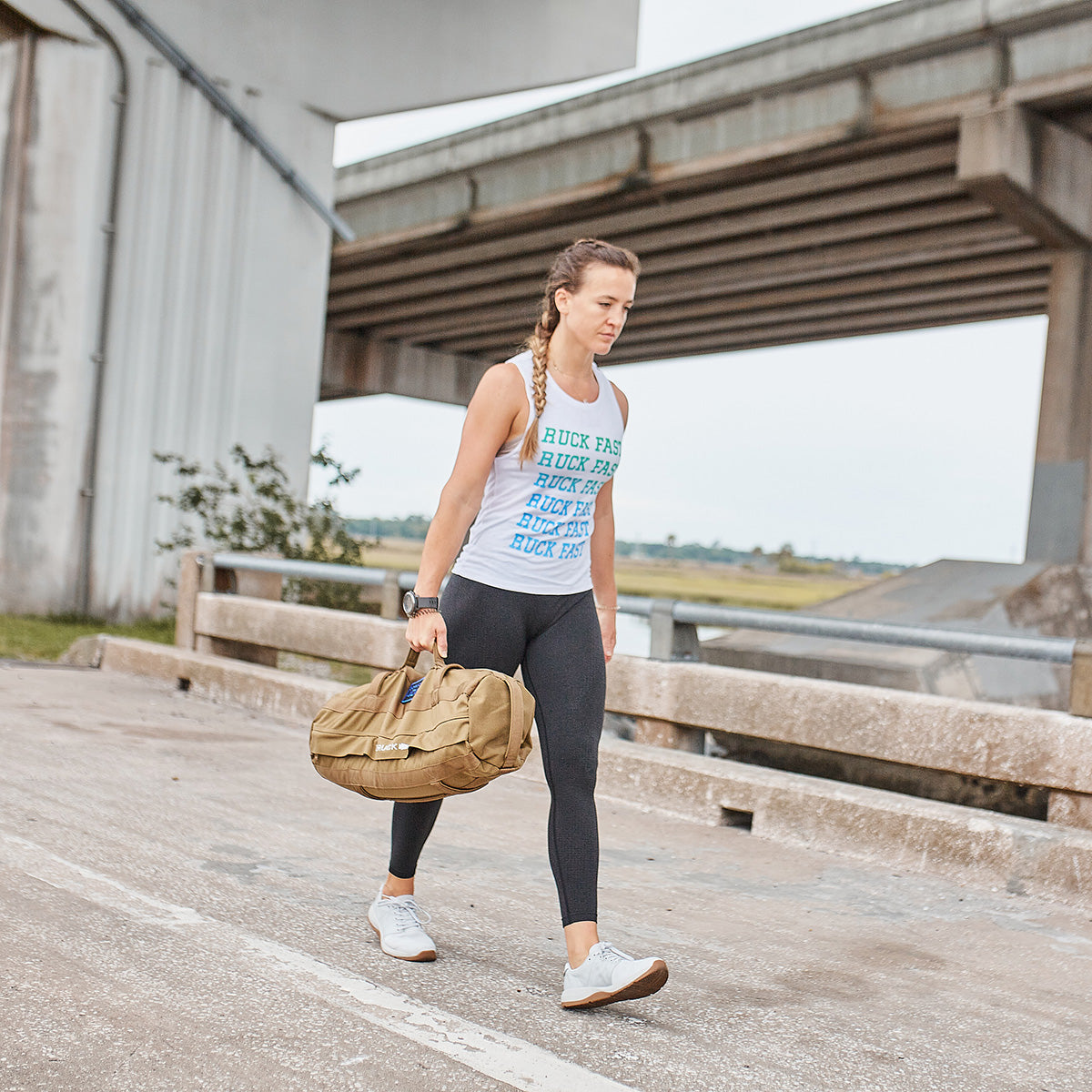 A woman with a long braid strides along a concrete path beneath a bridge, carrying a tan duffel bag as part of the GORUCK Teen/Preteen Ruck Training Program. She is dressed in a sleeveless shirt, leggings, and sneakers, with the background featuring a grassy field and the bridge structure.