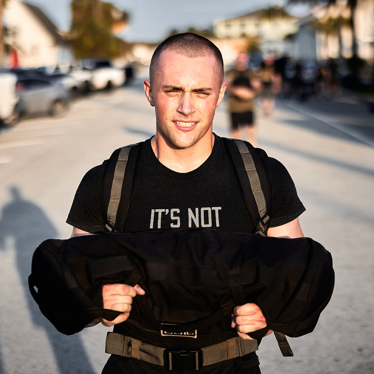 A person with a shaved head, clad in a black T-shirt, is taking part in GORUCK's Teen/Preteen Ruck Training Program by hoisting a sandbag over their shoulder. They confidently stride down the street surrounded by parked cars and passersby, proudly demonstrating their commitment to strength and conditioning.