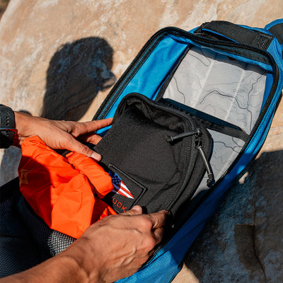 A person methodically packs gear into a blue duffel bag on the rock, including an orange jacket and a black pouch with a patch, while a GORUCK Bullet Ruck made of Ripstop ROBIC® nylon rests nearby.