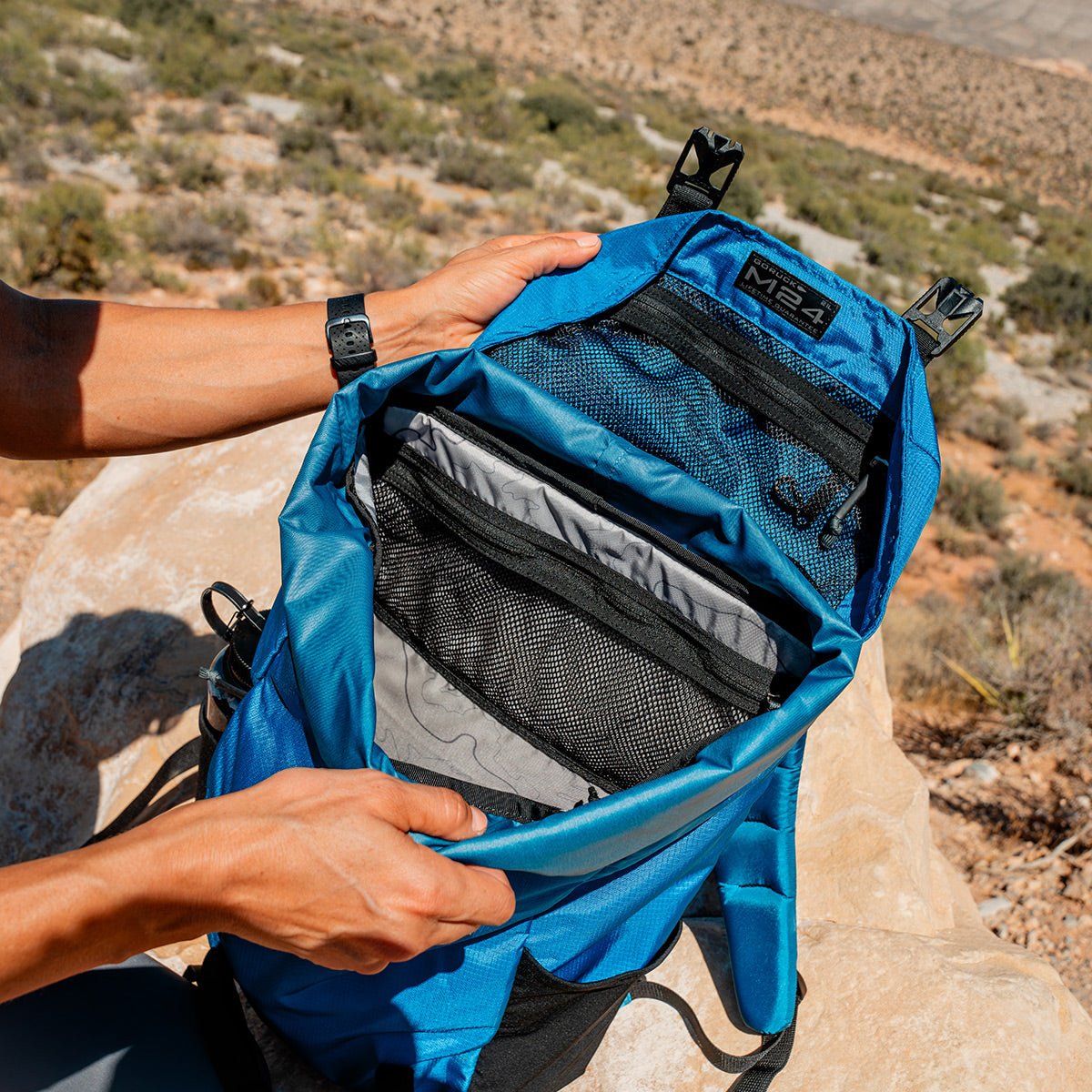 An individual outdoors holds an open GORUCK M24 - Ripstop ROBIC® rucksack on a rocky surface, featuring resilient materials and set against a desert backdrop.