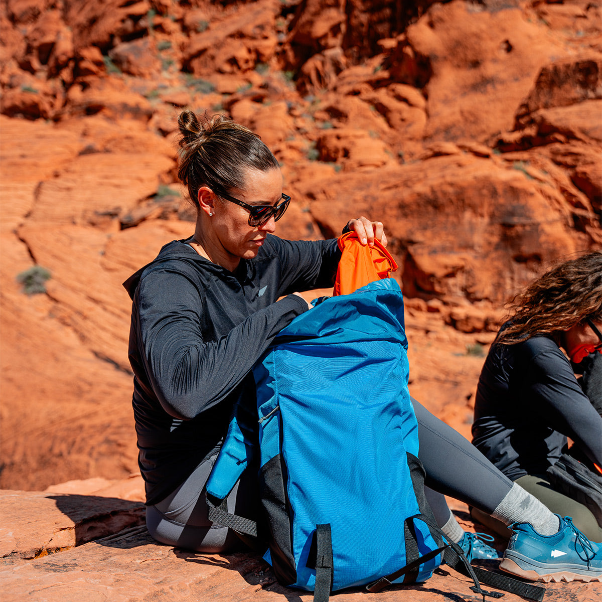 In the sunlit red desert landscape, a person sits on rocks, unpacking the GORUCK M24 rucksack crafted from durable Ripstop ROBIC® materials.