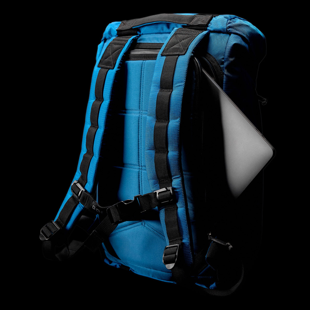 A GORUCK M24 - Ripstop ROBIC® rucksack, featuring a sleek blue design with black straps, is set against a dark background, showing a laptop partially exposed in its side compartment. Made from robust materials, this contemporary backpack is ideal for work or school.