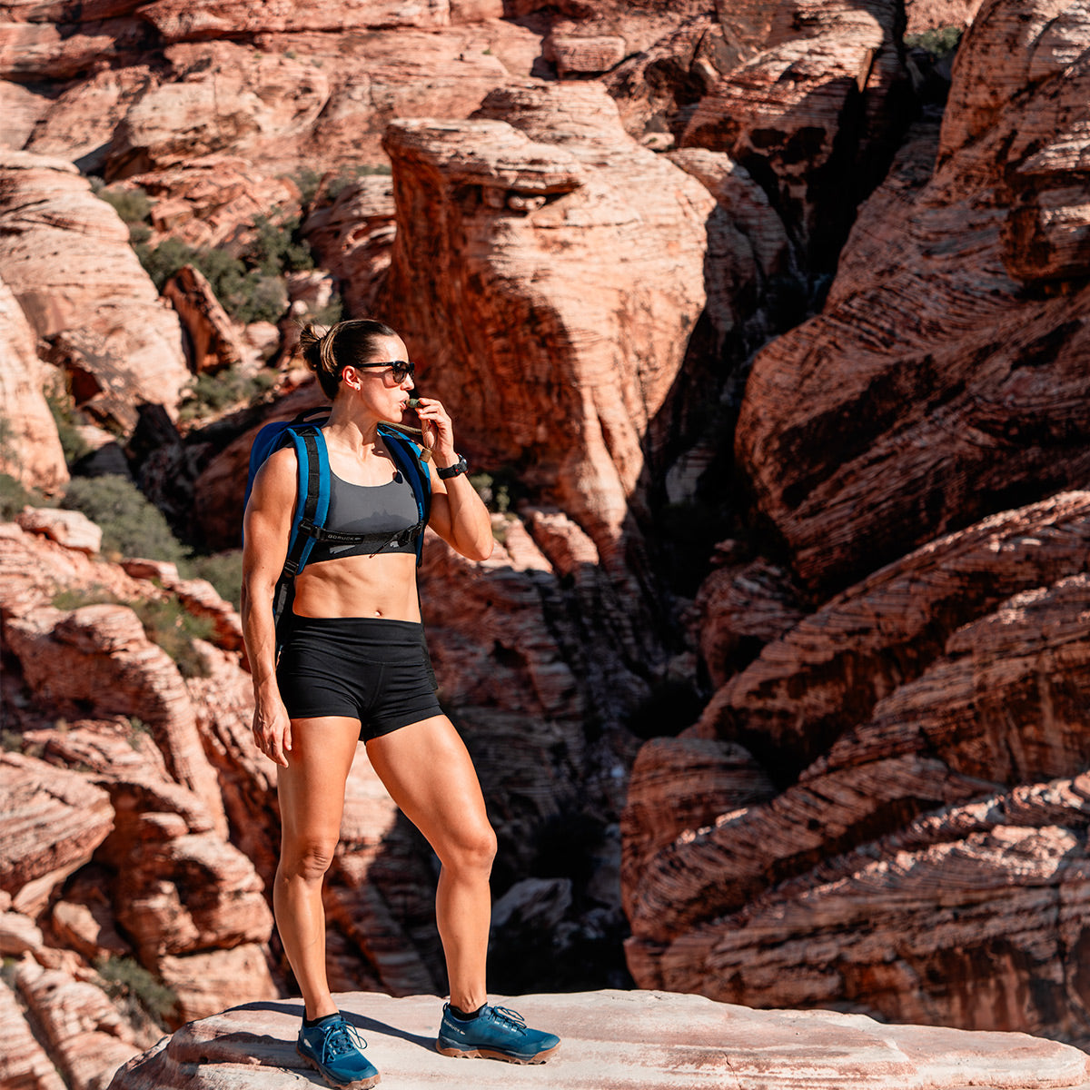 A woman in athletic gear stands confidently on a rock, her GORUCK Rucker 4.0 rucksack slung over one shoulder, surrounded by the breathtaking expanse of a red rock canyon landscape.