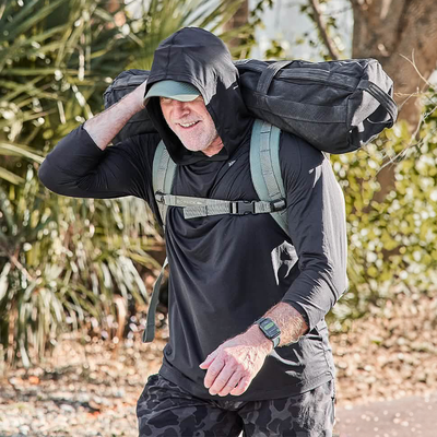 A person wearing the GORUCK Men's Sun Hoodie - ToughMesh in black strolls outdoors, smiling under UPF 30+ sun protection and carrying a large black bag over their shoulder. They sport a green watch and camo-patterned pants, moving effortlessly with featherweight performance amidst vibrant greenery.