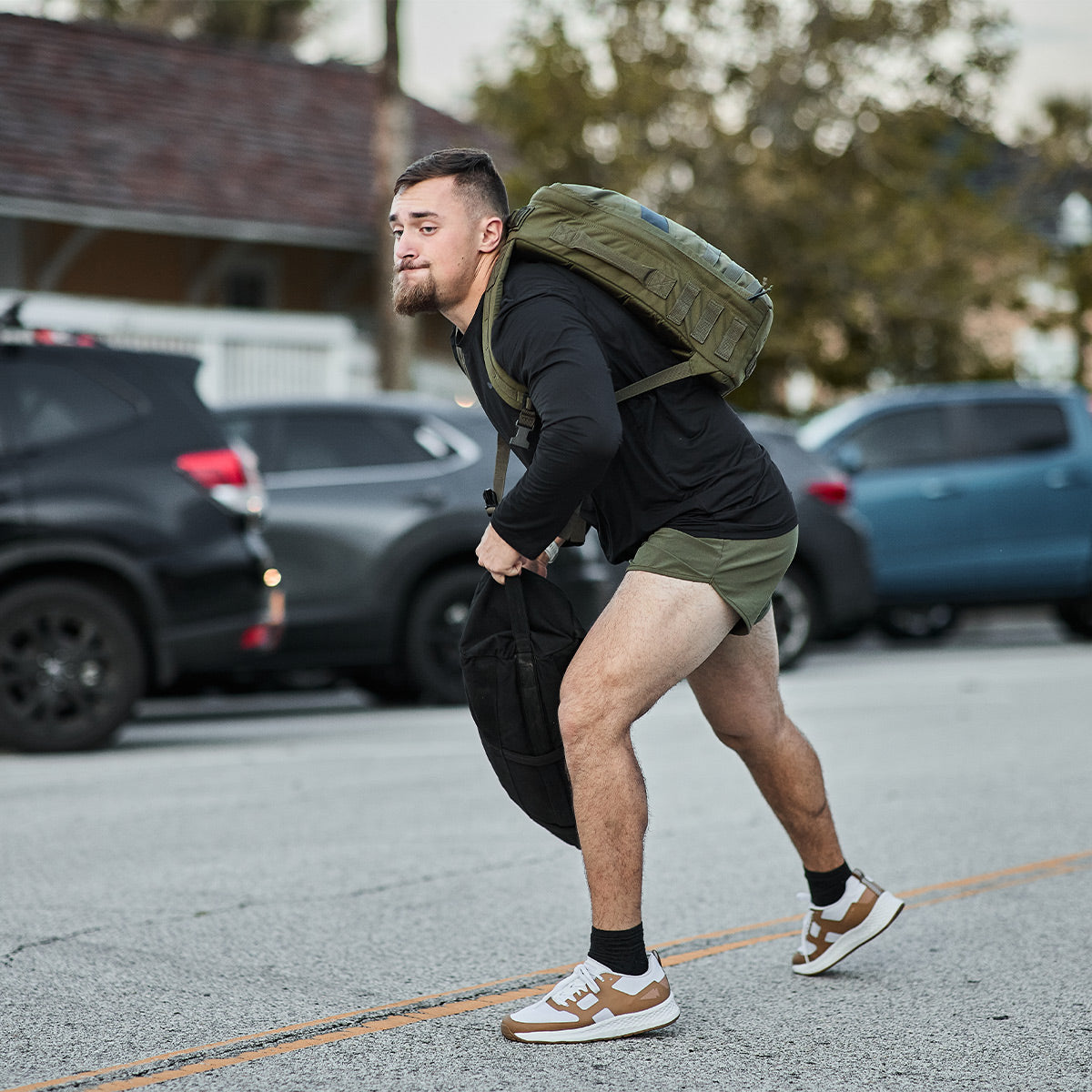 A man with a beard wearing a backpack and green shorts is bent over, holding a black bag crafted from GORUCK's ToughMesh™ fabric. He's on a street with parked cars and trees in the background, looking as if he's in a hurry.