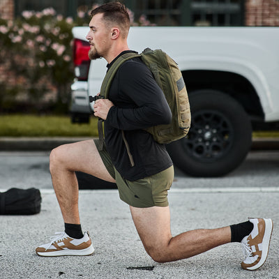 A man with a short haircut is exercising outdoors, performing a lunge while carrying an olive green backpack. He is wearing a black ToughMesh long sleeve performance tee from GORUCK, green shorts, black socks, and brown sneakers. A white truck and some greenery are visible in the background.