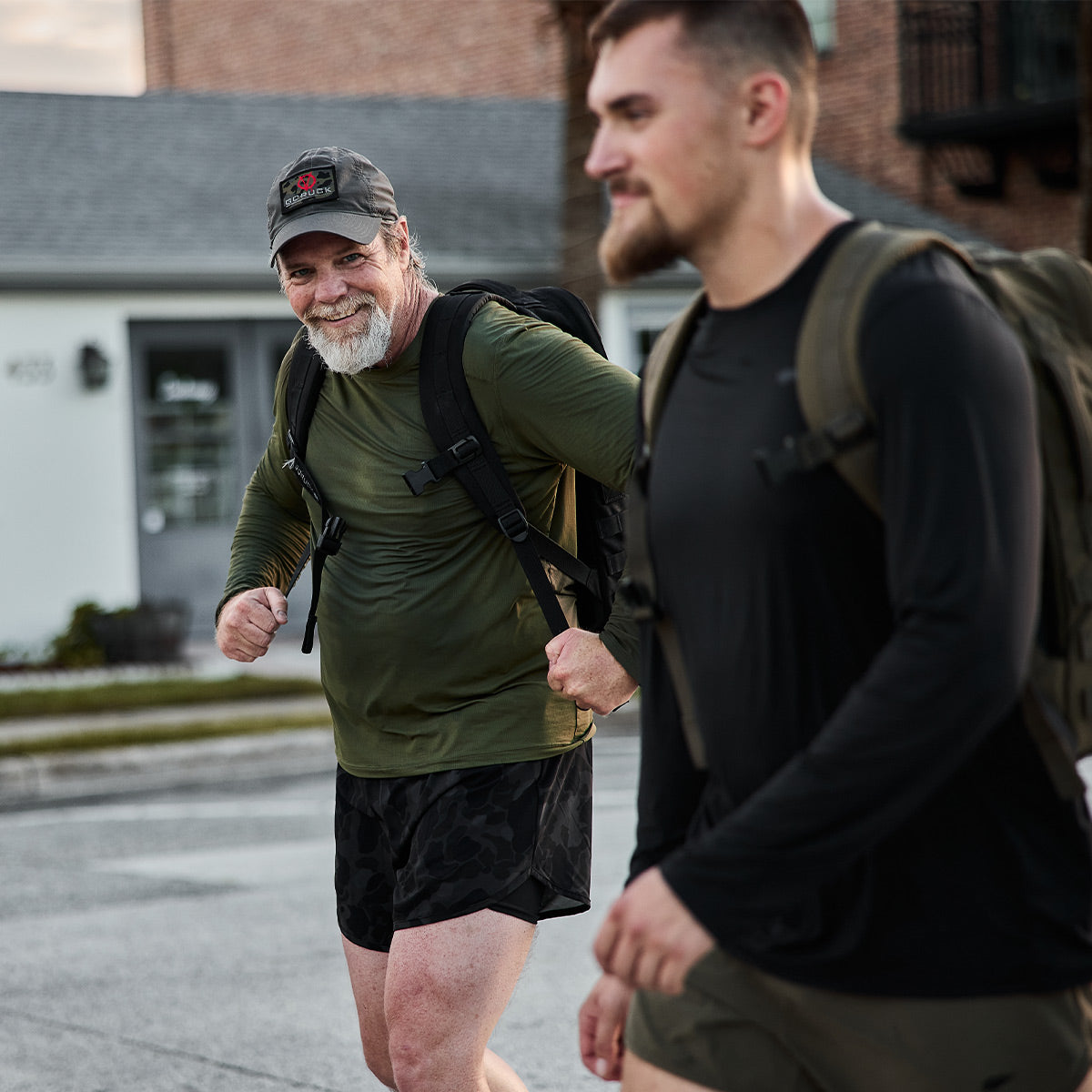 Two men strolling outside with backpacks, wearing GORUCK Men's Long Sleeve Performance Tees made from ToughMesh fabric and shorts. The older man on the left smiles while the younger peers ahead. They seem to chat casually with brick buildings behind them, perhaps envisioning an adventure in the Italian Alps.