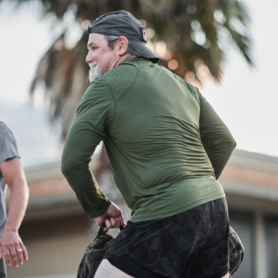 A man wearing the Men's Long Sleeve Performance Tee - ToughMesh by GORUCK and black shorts, with a backward cap, is participating in outdoor physical activity. He is lifting or moving an item with both hands, with a palm tree and building in the background.