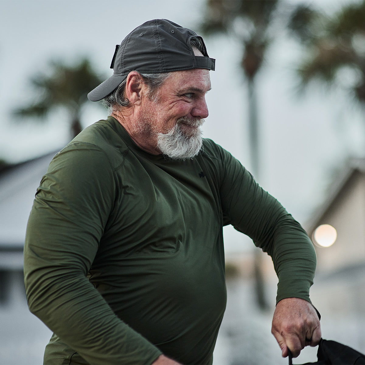 A man with a gray beard and a backward gray cap is smiling outdoors while wearing the Men's Long Sleeve Performance Tee - ToughMesh from GORUCK. Out-of-focus palm trees and buildings provide a backdrop reminiscent of the scenic Italian Alps.