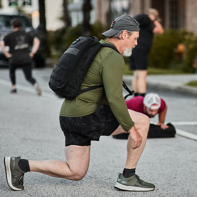 An older man wearing a GORUCK Men's Long Sleeve Performance Tee - ToughMesh, black shorts, and a backpack lunges on a street, exercising. The ToughMesh™ fabric of his tee offers maximum comfort. Other people in athletic apparel can be seen exercising in the background.