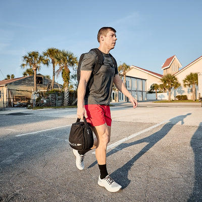 A person in a gray GORUCK Men’s Performance Tee - ToughMesh and red shorts is walking on a paved area, carrying a black sports bag. The background features palm trees, houses, and a clear blue sky, creating a serene contrast to an adventurous day in the Italian Alps.